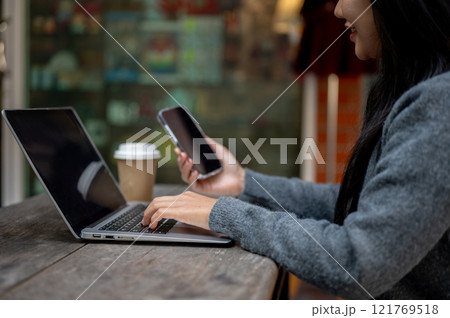 A woman holding her smartphone while working on her laptop, sitting at a table outdoors. 121769518