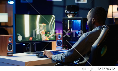 Man having fun by playing videogames on gaming PC at computer desk, enjoying day off from work. Gamer battling enemies in online multiplayer shooter from the comfort of his home, camera A 121770312