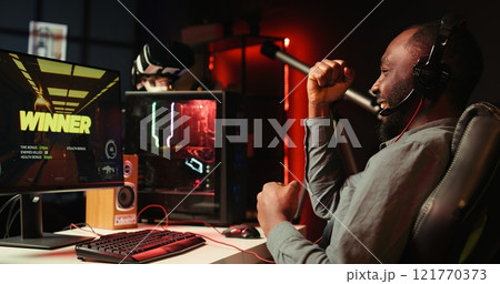 Man feeling happy while playing videogames on computer desktop with headphones on, celebrating victory. Cheerful gamer smiling after winning game on gaming PC while at home, camera B 121770373