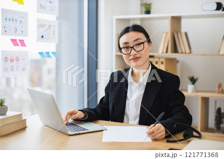 Young Asian businesswoman sitting at desk with laptop, taking notes. Professional setting includes bookshelves, diagrams on window. Elegant and focused workplace atmosphere. 121771368