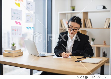 Young Asian businesswoman wearing glasses writing notes. Modern office setting with laptop, books, and plant. Concept of focus, productivity, and professional work environment. Young Asian businesswoman wearing glasses writing notes. Modern office setting with laptop, books, and plant. Concept of focus, productivity, and professional work environment. 121771376