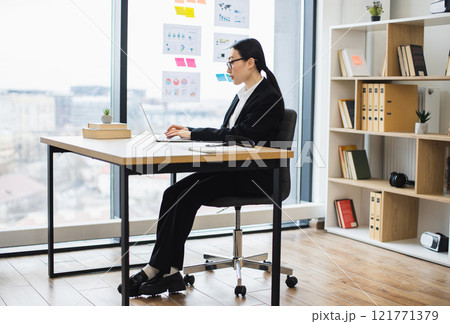 Asian businesswoman in professional attire working at laptop in modern office environment. Young adult focused on tasks, highlighting workplace productivity and focus. Asian businesswoman in professional attire working at laptop in modern office environment. Young adult focused on tasks, highlighting workplace productivity and focus. 121771379