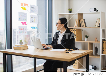 Asian businesswoman wearing glasses working on laptop at modern office desk. Professional atmosphere with natural light and organized workspace. Focused and productive environment. Asian businesswoman wearing glasses working on laptop at modern office desk. Professional atmosphere with natural light and organized workspace. Focused and productive environment. 121771383