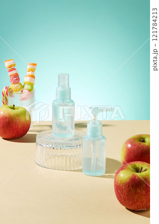 A cosmetics advertisement set against a light blue backdrop. An apple and some sweets in a glass, a blue mockup of cosmetics placed on a glass podium, on a yellow tabletop. 121784213