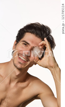Smiling, handsome, brunette man places ice cube under eye with gentle touch, during refreshment skincare process, against white background. Smiling, handsome, brunette man places ice cube under eye with gentle touch, during refreshment skincare process, against white background. 121785114