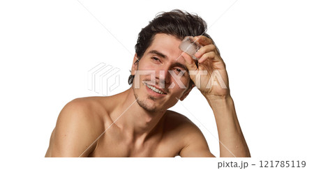 Close-up of shirtless man smiling, holding ice cube over eyebrow against white background. Clear skin and joyful mood. 121785119