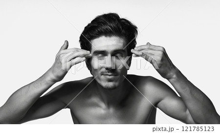 Black-and-white portrait of shirtless man applies cream to his face, enjoying its moisturizing effect against white background. Black-and-white portrait of shirtless man applies cream to his face, enjoying its moisturizing effect against white background. 121785123