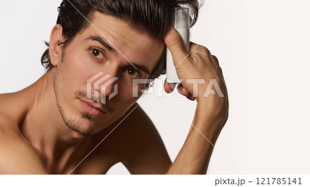 Close up photo of handsome, smiling man sitting in casual pose and holding battle of cosmetic product for facial care against white background. Close up photo of handsome, smiling man sitting in casual pose and holding battle of cosmetic product for facial care against white background. 121785141