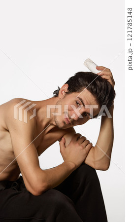 Stylish young man seated in relaxed pose, resting his head on his hand while holding bottle of cream, against white background. 121785148