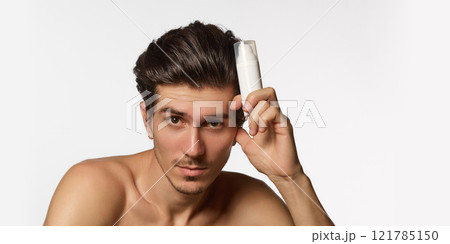 Confident, and direct man, with styled hair and clean smooth skin, holding white bottle near his head, looking at camera against white background. Confident, and direct man, with styled hair and clean smooth skin, holding white bottle near his head, looking at camera against white background. 121785150