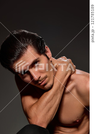 Man tilting his head slightly, with one arm resting on his shoulder. His confident gaze and sleek hairstyle emphasized against dark background. Man tilting his head slightly, with one arm resting on his shoulder. His confident gaze and sleek hairstyle emphasized against dark background. 121785189