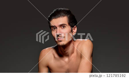 Shirtless man with slicked-back hair and stubble, gazes directly at camera, displaying confident expression against deep dark background. Shirtless man with slicked-back hair and stubble, gazes directly at camera, displaying confident expression against deep dark background. 121785192