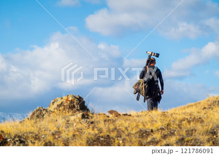 Photographer hiking with camera gear through grassy landscape under blue sky 121786091