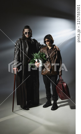 Vampire and companion posing in stylish outfits, female companion holds potted plant, leather bag in other hand. Vampire and companion posing in stylish outfits, female companion holds potted plant, leather bag in other hand. 121788301
