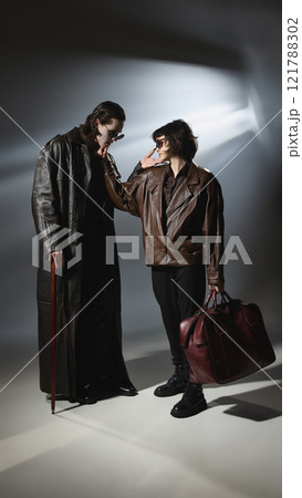 Vampire leans toward female companion in brown jacket, looks on her with love. Spotlight casts shadows on models in stylish outfits. Vampire leans toward female companion in brown jacket, looks on her with love. Spotlight casts shadows on models in stylish outfits. 121788302