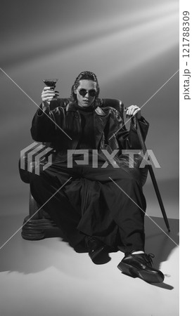 Black-and-white photo of man in a leather coat and sunglasses, looks as vampire, sitting in chair and toasting holding cocktail. Party mood. Black-and-white photo of man in a leather coat and sunglasses, looks as vampire, sitting in chair and toasting holding cocktail. Party mood. 121788309