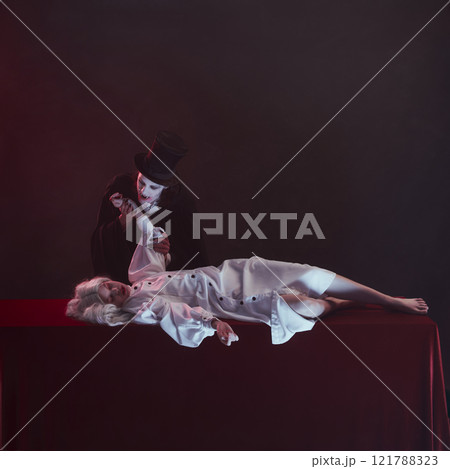 Vampire leans over woman dressed in white lying on table with red cloth, holding their wrist as if about to bite, with intense and haunting expressions. 121788323