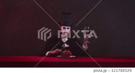 Vampire sits at table with red cloth, wearing top hat and black cloak, holding glass of red wine in one hand and heart on plate in front. 121788329