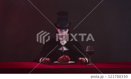 Terrifying man, vampire in cape and top hat sits at red table with glass of wine and eating heart on plate, look forward with serious expression. 121788330