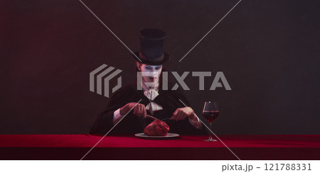 Terrifying man, vampire in cape and top hat sits at red table with glass of wine and heart on plate, staring forward with serious expression. 121788331