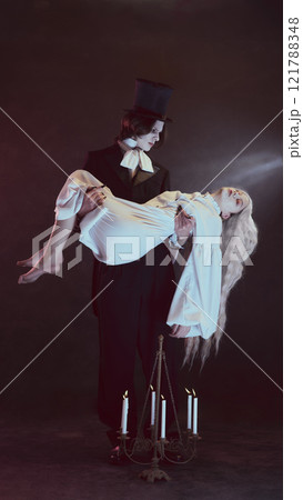 Vampire, in formal black suit carries woman in flowing white dress, looks as ghost, with lit candles visible below them. 121788348