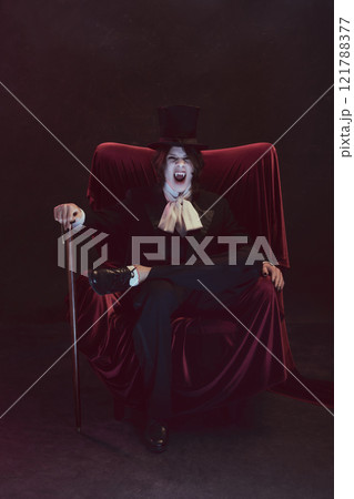Angry man with white face in formal suit, looks as vampire, sits on velvet chair with cane against dark background. 121788377
