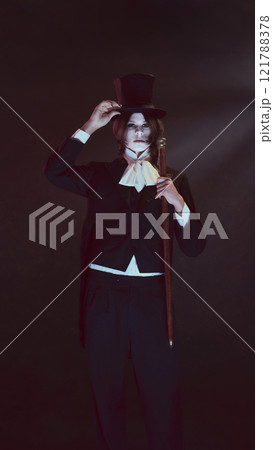 Pale-faced man, vampire, dressed in sleek black attire and top hat stands holding cane, looking serious under dim light. Pale-faced man, vampire, dressed in sleek black attire and top hat stands holding cane, looking serious under dim light. 121788378