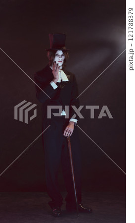 Pale-faced man, vampire, dressed in sleek black suit and top hat stands holding cane, looking serious under dim light. 121788379