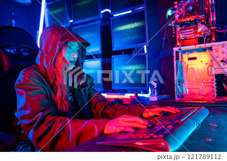 Woman hacker. Asian woman wearing hood is typing keyboard for coding hacking programming system. room is dimly lit. possibly typing or browsing internet, Hacker in neon light, Female gamer play game Woman hacker. Asian woman wearing hood is typing keyboard for coding hacking programming system. room is dimly lit. possibly typing or browsing internet, Hacker in neon light, Female gamer play game 121789112