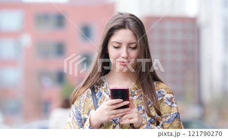 Beautiful woman with long blond hair holding mobile phone, looking at mobile phone screen, using smartphone apps. posing on street. Positive model smiling. She is standing with smartphone in hands on Beautiful woman with long blond hair holding mobile phone, looking at mobile phone screen, using smartphone apps. posing on street. Positive model smiling. She is standing with smartphone in hands on 121790270