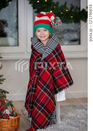 Happy little girl wearing santa claus hat wrapped in warm checkered blanket in christmas room. Happy little girl wearing santa claus hat wrapped in warm checkered blanket in christmas room. 121790658