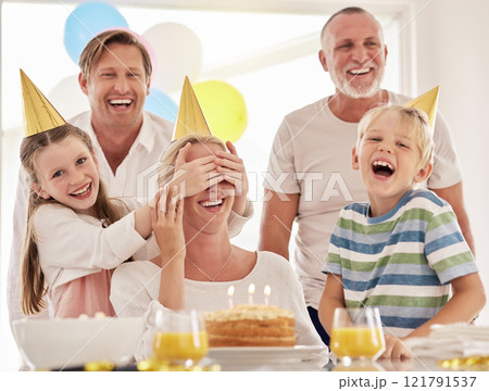 Little girl covering mothers eyes while celebrating her birthday or surprising her on Mothers day. Woman having a party at home with her family while wearing party hats and having fun together Little girl covering mothers eyes while celebrating her birthday or surprising her on Mothers day. Woman having a party at home with her family while wearing party hats and having fun together 121791537