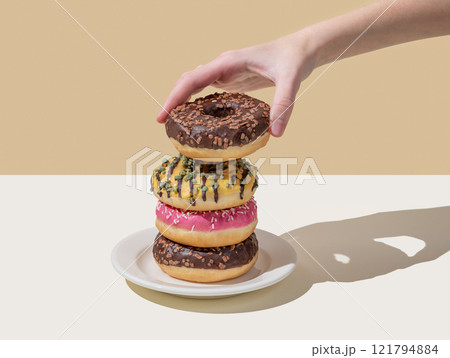 Hand takes one donut from stack of glazed donuts with different toppings on beige white background. 121794884