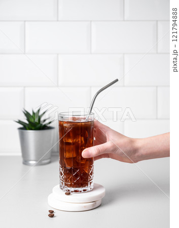 Hand holding glass of cold brew coffee with ice and straw on light table against brick background Hand holding glass of cold brew coffee with ice and straw on light table against brick background 121794888