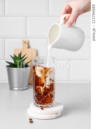 Cold latte coffee in a tall glass with hand pouring alternative milk from a milk jug on light table Cold latte coffee in a tall glass with hand pouring alternative milk from a milk jug on light table 121794889