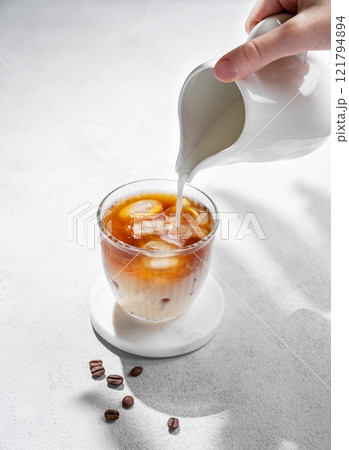 Cold latte coffee in a glass with hand pouring alternative milk from a milk jug on light background Cold latte coffee in a glass with hand pouring alternative milk from a milk jug on light background 121794894