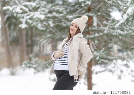 Young pregnant woman in a snowy winter forest with copy space. Expecting mother holding her belly and smiling, outdoors. Concept of maternity, love and joy of pregnancy surrounded by nature. 121798082