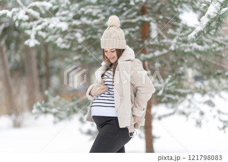 Happy young pregnant woman in a snowy winter forest. Expecting mother smiling and looking at her belly outdoors, natural light. Concept of maternity, love and joy of pregnancy surrounded by nature. 121798083