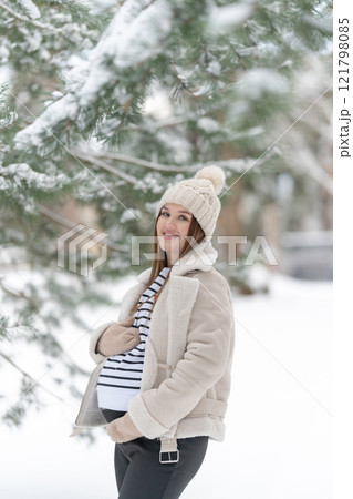 Cheerful young pregnant woman in a snowy winter forest with space for text. Expecting mother smiling and holding her belly outdoors. Concept of maternity, love, waiting, and the joy of pregnancy. 121798085