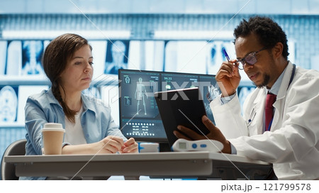 African american medic presenting the diagnostic reports to a female patient, showing the lab test results after medical examination. Physician offering professional advice. Camera A. 121799578