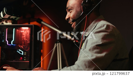 Smiling gamer with headphones on relaxing in gaming room, playing videogames on powerful computer, chatting with friends. Cheerful esports player performing in gaming tournament, camera B Smiling gamer with headphones on relaxing in gaming room, playing videogames on powerful computer, chatting with friends. Cheerful esports player performing in gaming tournament, camera B 121799637