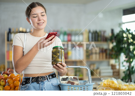 European woman customer at shop scans QR code on pickled cucumbers jar using phone camera 121803042