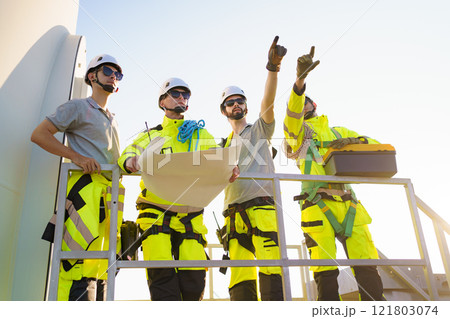 Team of professional electrical engineers in fully safety suit are working and discussing together at the windmill electric generating turbine. Electricians working at the site of windmill turbine. 121803074