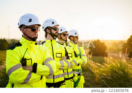Group portrait of electrical engineers team pose to camera against the sunset nature in background. Engineers in fully safety suit and headwear are posing to camera. 121803076