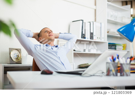 Relaxed office worker sitting at workplace with hands behind head 121803369