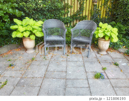 Calm serene and inviting yard is for people to relax unwind and enjoy peaceful area. Outdoor garden with grey chairs potted plants greenery around and grey tiled floors on sunny day Calm serene and inviting yard is for people to relax unwind and enjoy peaceful area. Outdoor garden with grey chairs potted plants greenery around and grey tiled floors on sunny day 121806193