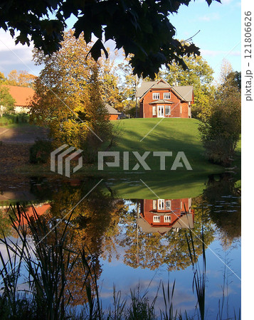 A small red house on a green hill is reflected in the mirror surface of the pond 121806626