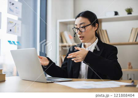 Asian businesswoman in headset conducting video call on laptop in modern office. Woman wearing glasses and suit appears professional and focused, engaging in conversation. 121808404