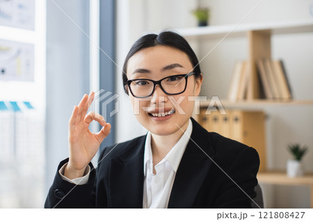 Asian businesswoman wearing glasses showing ok sign, smiling confidently in office setting. Captures positive energy, professional attire, and modern work environment, conveying success and approval. 121808427