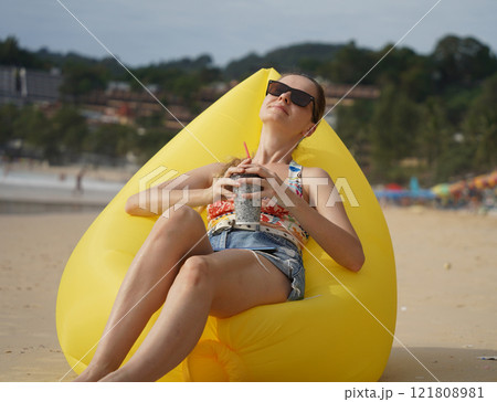A young woman on the beach lounging on a yellow bean bag  121808981